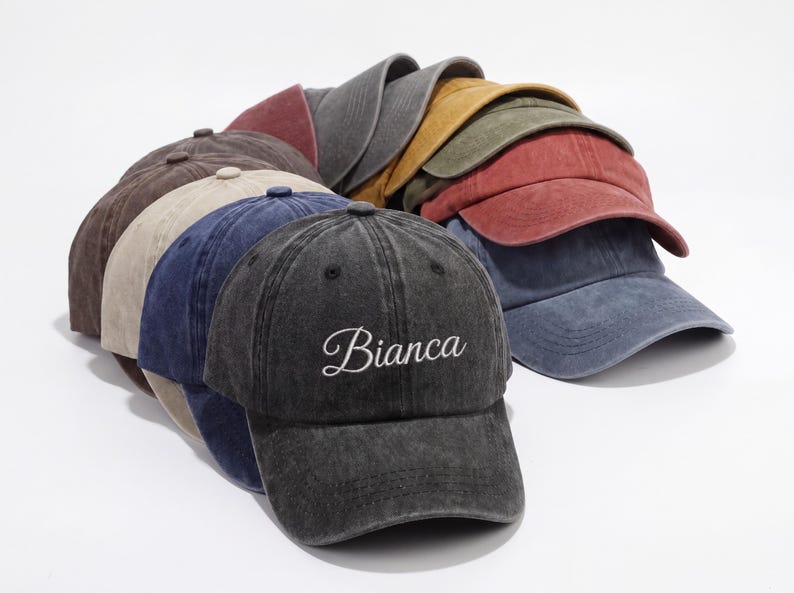 May include: A stack of baseball caps in various colors, including gray, brown, blue, and red. The front cap is dark gray with the name "Bianca" embroidered in white script. The caps have a worn, vintage look.