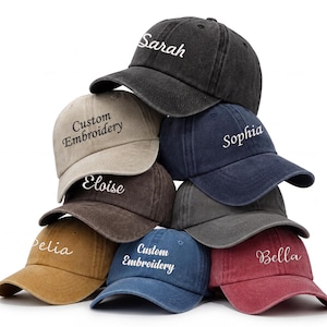 May include: A stack of baseball caps in various colors, including black, tan, navy, and red. Each cap features custom embroidery with names or the phrase "Custom Embroidery" in white script. The caps have a classic, worn look.