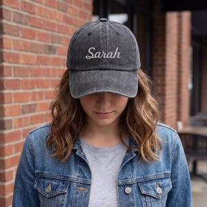 May include: A gray baseball cap with the name "Sarah" embroidered in white script. The hat is worn by a person wearing a denim jacket and a gray t-shirt. The background includes a brick wall and outdoor seating.