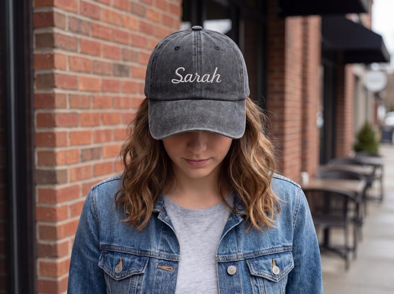 May include: A gray baseball cap with the name "Sarah" embroidered in white script. The hat is worn by a person wearing a denim jacket and a gray t-shirt. The background includes a brick wall and outdoor seating.