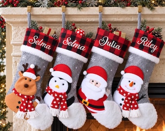 May include: Four gray felt Christmas stockings with white faux fur cuffs and red and black buffalo plaid tops. Each stocking features a personalized name embroidered in white script and a plush character: a reindeer, penguin, Santa, and snowman.