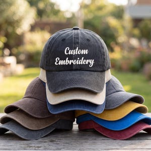 May include: A stack of baseball caps in various colors, including black, brown, beige, blue, yellow, and burgundy. The top cap is black with the words "Custom Embroidery" embroidered in white.