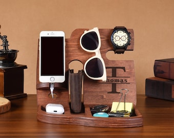May include: A dark brown wooden desk organizer with compartments for a phone, wallet, sunglasses, watch, and small accessories. The organizer is engraved with the name "Thomas". A white phone is charging.