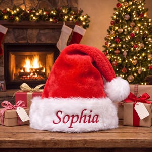 Può includere: Un cappello da Babbo Natale in peluche rosso con bordo in pelliccia sintetica bianca e il nome Sophia ricamato in corsivo rosso. Il cappello è posizionato su una superficie in legno tra due pacchetti regalo con nastri ed etichette vuote.