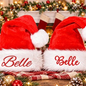 May include: Two bright red plush Santa hats with white faux fur trim. Each hat features the name Belle embroidered in red script on the front white band. The hats have a classic pointed shape with a white pom-pom at the tip of each.