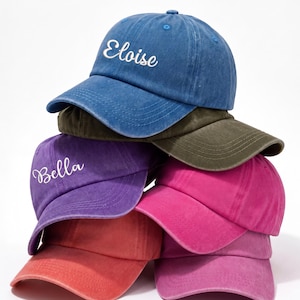 May include: A stack of six colorful baseball caps. The top cap is blue with the name "Eloise" embroidered in white. Other caps are purple, pink, red, and olive green. The caps are made of a soft material and have a curved brim.