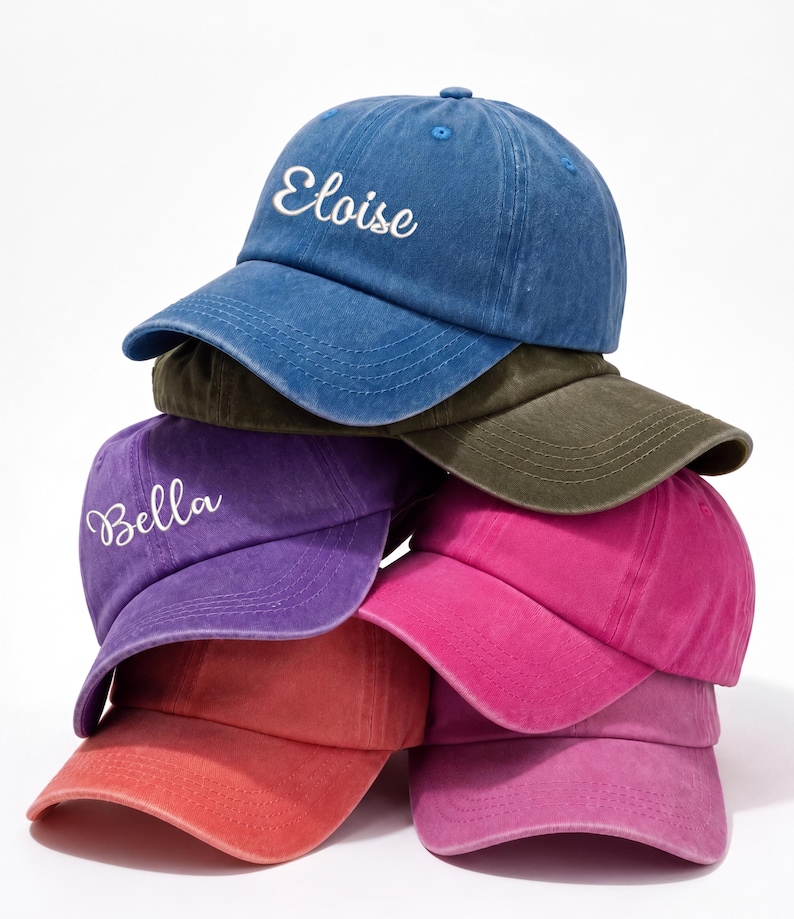 May include: A stack of six colorful baseball caps. The top cap is blue with the name "Eloise" embroidered in white. Other caps are purple, pink, red, and olive green. The caps are made of a soft material and have a curved brim.