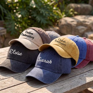 May include: A collection of custom embroidered baseball caps in various colors, including black, tan, gold, blue, and maroon. Each cap features personalized text, with names like "Eloise" and "Sarah" visible. The caps are arranged on a wooden surface.