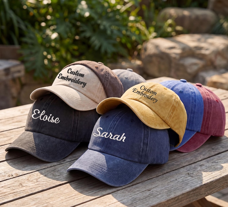 May include: A collection of custom embroidered baseball caps in various colors, including black, tan, gold, blue, and maroon. Each cap features personalized text, with names like "Eloise" and "Sarah" visible. The caps are arranged on a wooden surface.