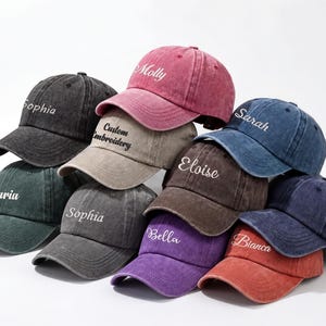 May include: A collection of baseball caps in various colors, including black, green, pink, and blue. Each cap features a curved brim and embroidered text with names like "Sophia" and "Molly". The caps are made of a worn, vintage-style fabric.