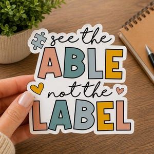 Może przedstawiać: Wykrawana naklejka z napisem see the ABLE not the LABEL wykonanym z różowych, niebieskich i żółtych liter blokowych. Projekt zawiera małą szarą ikonę puzzla oraz dwa serca, jedno żółte i jedno różowe, na białym tle.