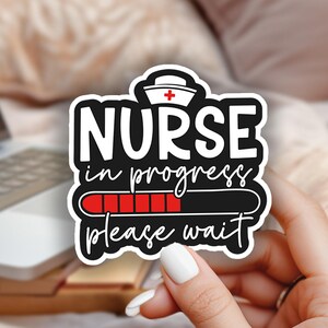 May include: A white sticker with the text "NURSE in progress please wait" and a red progress bar. The sticker is held by a hand with white nail polish. A laptop and pink flowers are in the background.
