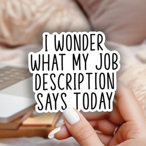 May include: A white sticker with black text that reads "I WONDER WHAT MY JOB DESCRIPTION SAYS TODAY" is held in a hand. A laptop, flowers in a jar, and a wooden tray are in the background.