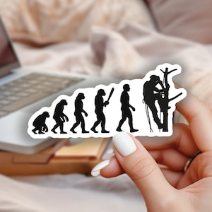 May include: A black and white sticker depicting the evolution of man, culminating in a tree climber. The sticker is held in a hand with white nail polish. A laptop and flowers are in the background.
