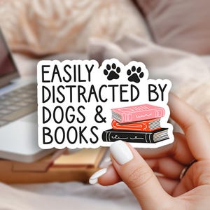 May include: A white sticker with the text "EASILY DISTRACTED BY DOGS & BOOKS" and paw prints. The sticker also features illustrations of stacked books in pink, orange, and black. The background includes a laptop, flowers, and a soft, neutral-toned setting.