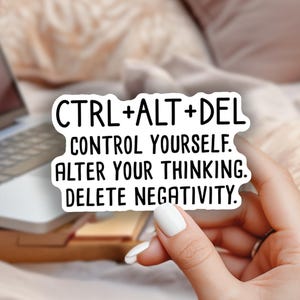 May include: A white sticker with the text "CTRL+ALT+DEL CONTROL YOURSELF. ALTER YOUR THINKING. DELETE NEGATIVITY." is held in a hand. A laptop, flowers, and a wooden tray are in the background.