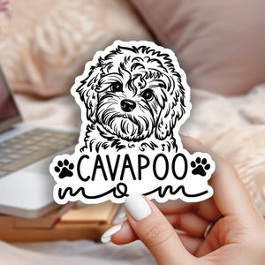 Pode incluir: Um autocolante a preto e branco com uma ilustração de um cão Cavapoo. O autocolante tem o texto "CAVAPOO mom" num estilo manuscrito, com pegadas. O autocolante é segurado por uma pessoa. Computador portátil e flores no fundo.