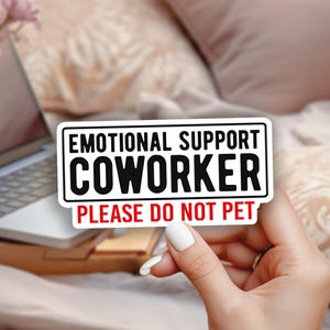 May include: A white sticker with black text that reads "EMOTIONAL SUPPORT COWORKER PLEASE DO NOT PET" is held in front of a laptop and a vase of pink flowers. The background includes a bed and pillows.