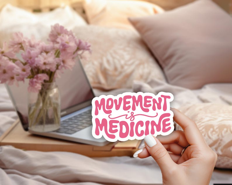 May include: A pink and white sticker with the words "MOVEMENT is MEDICINE" in a playful font. The sticker is held in front of a laptop, flowers, and pillows.