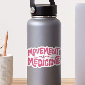 May include: A gray water bottle with a black lid and handle. The bottle features a pink sticker with the words "MOVEMENT is MEDICINE" in a playful, bubble-style font. A pencil and a white cup are visible in the background.