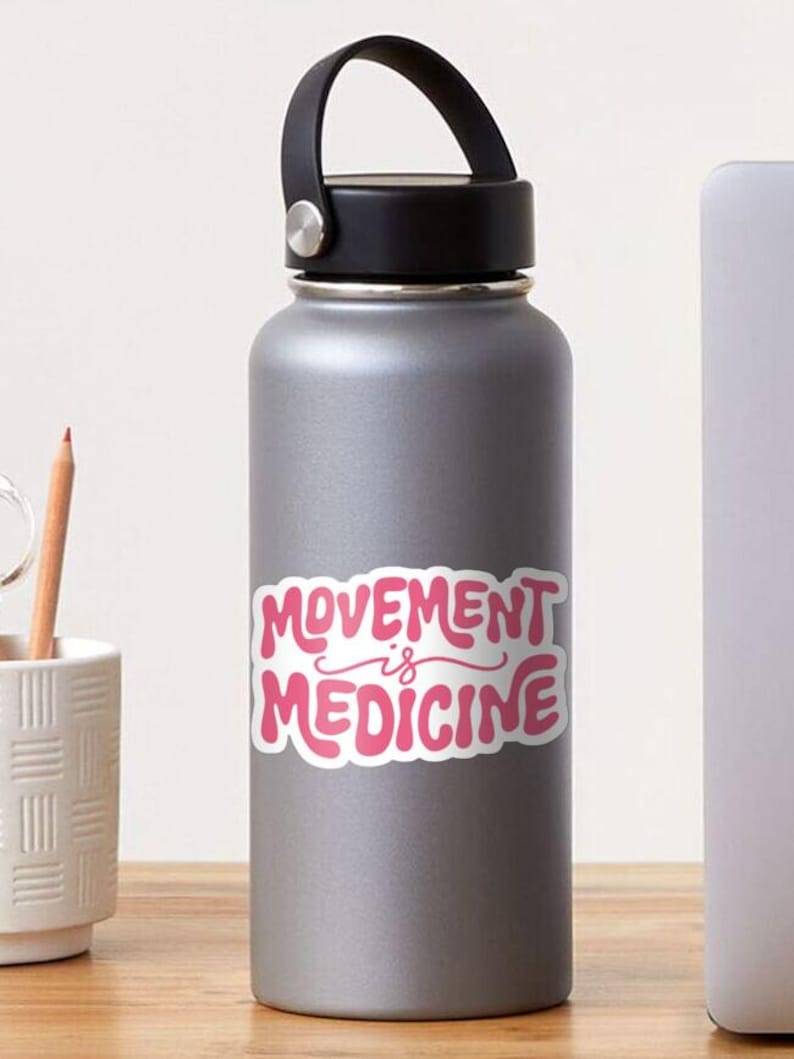 May include: A gray water bottle with a black lid and handle. The bottle features a pink sticker with the words "MOVEMENT is MEDICINE" in a playful, bubble-style font. A pencil and a white cup are visible in the background.