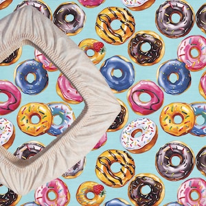 May include: A light beige fitted sheet with a pattern of colorful donuts on a light blue background. The donuts have various toppings, including chocolate, sprinkles, and fruit.