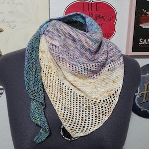 May include: A triangular shawl with a crocheted border. The shawl is made with a gradient yarn that transitions from teal to purple to cream. The crocheted border is cream and has a lacy pattern.