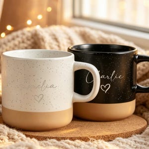 Könnte beinhalten: Zwei Keramikbecher, einer in Weiß und einer in Schwarz, beide mit gesprenkeltem Design im oberen Bereich und einem einfarbigen beigen Boden. Jeder Becher ist mit einem Namen in Schreibschrift und einem kleinen Herzsymbol auf der Vorderseite versehen.