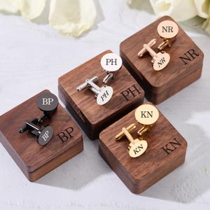 May include: A collection of personalized cufflinks in various metallic finishes, including silver, gold, and black. Each pair rests on a wooden box, engraved with initials. The cufflinks feature a circular or oval design with engraved initials, perfect for a gift.