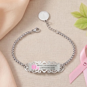 Puede incluir: Una pulsera de alerta médica en tono plateado con una cadena de eslabones cuadrados y una placa rectangular decorativa. La placa está grabada con el texto BREAST CANCER SURVIVOR, LYMPHEDEMA ALERT y NO BP/IV/NEEDLES THIS ARM, junto a un lazo rosa.