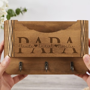 May include: A wooden key holder with the word "PAPA" engraved in large letters. Below, names Claude, Daniel, and Rosalie are also engraved. The holder has three hooks and a slot for mail. The wood has a natural finish.