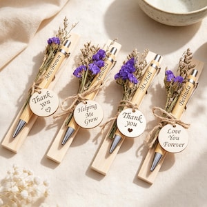 Puede incluir: Cuatro bolígrafos de bambú con puntas plateadas, sujetos a una base de madera con flores secas moradas y una etiqueta redonda de madera. Las etiquetas tienen grabado: Thank you, Helping Me Grow, Thank you, Love You Forever, junto a los nombres Mrs. Brown, Amanda, Ms. Hall, Belinda.