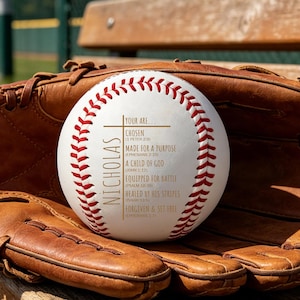 Puede incluir: Una pelota de béisbol blanca con costuras rojas y el nombre "Nicholas" impreso verticalmente. La pelota descansa en un guante de béisbol de cuero marrón. El texto en la pelota incluye "Elegido" y "Hecho para un propósito".