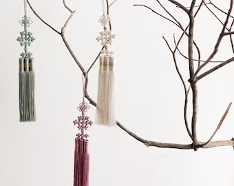 May include: Three decorative hanging ornaments featuring intricate knotted cord designs in sage green, off-white, and dusty rose. Each ornament includes a long, silky tassel with gold-toned metallic bands at the top, suspended by a thin matching cord.