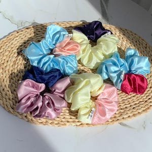 以下が含まれることがあります： 青、ピンク、黄色、紫など、さまざまな色のサテンシュシュのコレクション。 シュシュは、織られた円形のマットの上に配置されています。 いくつかのシュシュには、小さな白いタグが付いています。