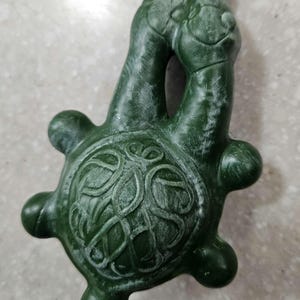 Pode incluir: Um pingente verde escuro em forma de tartaruga com uma argola na parte superior. O pingente apresenta um design detalhado na carapaça e duas cabeças de tartaruga voltadas uma para a outra. O pingente é feito de um material sólido, possivelmente esculpido.