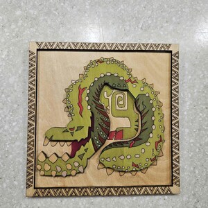 May include: A wooden square featuring a green and red monster design. The monster has a spiky exterior and a detailed interior. The border is a repeating triangle pattern. The background is a light tan color.