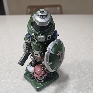 May include: A detailed figurine of a character in green armour, holding a gun and a shield with a red symbol. The figurine stands on a green base with a red skull. The character's helmet is silver and green.