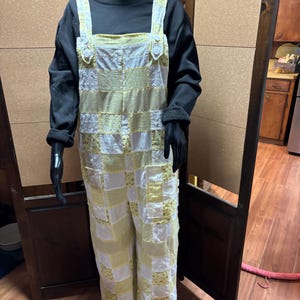 May include: A patchwork jumpsuit in white and yellow floral and solid fabrics. The jumpsuit has wide legs and adjustable straps. It is paired with a black long-sleeved shirt. The outfit is displayed on a mannequin.