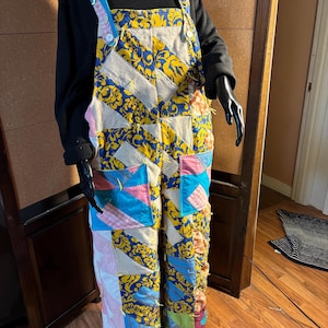 May include: Overalls with a patchwork design. The overalls feature a mix of fabrics in various colors, including blue, yellow, pink, and white. The bib and straps are made of a light blue and pink striped fabric. The overalls are displayed on a mannequin.