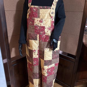 May include: Quilted patchwork overalls in a mix of floral and paisley patterns, featuring shades of red, cream, and brown. The bib overalls have adjustable straps and pockets. The garment is displayed on a mannequin.