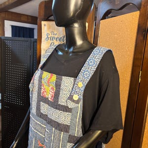 May include: A patchwork jumpsuit in shades of blue, with floral and geometric patterns. The jumpsuit has wide straps with a blue and white floral design, wooden buttons, and pockets. Worn over a black t-shirt.