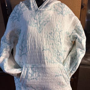 May include: A white quilted hoodie featuring a light blue coral reef pattern throughout. The garment includes long sleeves, a hood, and a large front kangaroo pocket. The fabric has a textured, grid-like stitched pattern across the entire surface.
