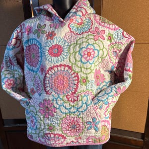 May include: A quilted hooded sweatshirt featuring a white base with a vibrant floral pattern in shades of pink, blue, and green. The garment includes long sleeves, a hood, and a large front kangaroo pocket, all constructed from a textured fabric.