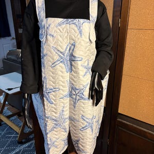 May include: White quilted overall shorts with a blue starfish pattern. The shorts have adjustable straps and pockets. The garment is displayed on a black mannequin.
