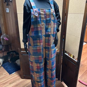 May include: Overalls with a patchwork design in shades of blue, red, and brown. The overalls have large front pockets and adjustable straps with button closures. The model is wearing a black long-sleeve shirt underneath.