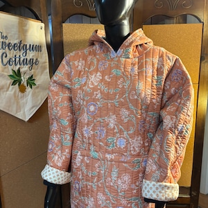 May include: A quilted orange hoodie with a floral pattern in blue, green, and white. The cuffs are folded back to reveal a white and cream geometric print. The hoodie is displayed on a black mannequin.