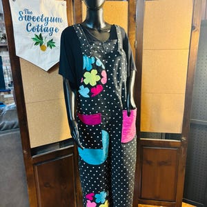 May include: Black jumpsuit with white polka dots, colorful floral and geometric patches, and pink pockets. The jumpsuit is worn over a black top. A banner with the text "The Sweetgum Cottage" and a pineapple design hangs in the background.