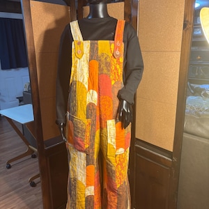 May include: Quilted jumpsuit in a patchwork design featuring shades of yellow, orange, and brown. The jumpsuit has a bib-style top with adjustable straps and large front pockets. The garment is displayed on a black mannequin.