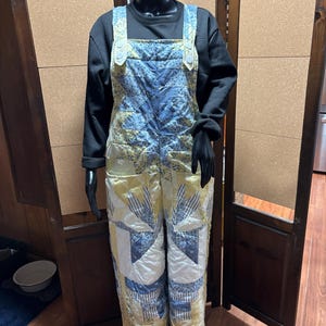 May include: A quilted overall with a patchwork design in shades of blue, yellow, and white. The overall is displayed on a mannequin, showcasing the intricate patterns and textures of the fabric. The overall has adjustable straps and a classic overall silhouette.